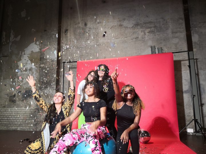 Fünf Mädchen und Frauen positionieren sich lachend zum Gruppenfoto. Dabei fliegt Konfetti. Im Hintergrund ist eine Leinwand und eine auf dem Boden liegende Diskokugel. Ein Mädchen sitzt auf einem Gymnastikball, neben ihr hockt links und rechts jeweils eine weitere Person. Zwei Projektteilnehmerinnen stehen mit aneinander gelehnten Köpfen dahinter.